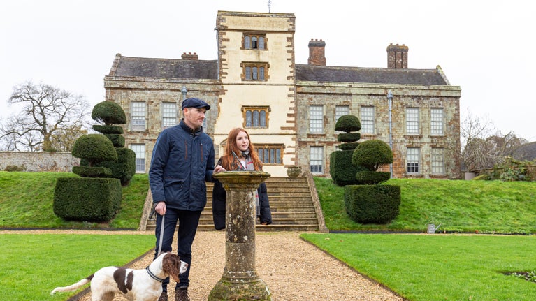 A man and young girl stand in the gardens at Canons Ashby. They are near a sundial with the house in the background. They have a dog on a lead.